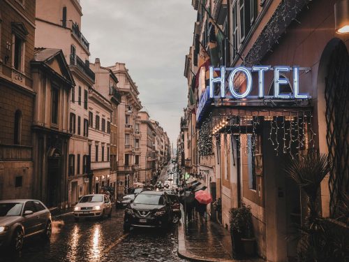 pexels-photo-2467558-2467558 A rainy city street scene at dusk, featuring a glowing neon hotel sign, wet cobblestones, and bustling cars.